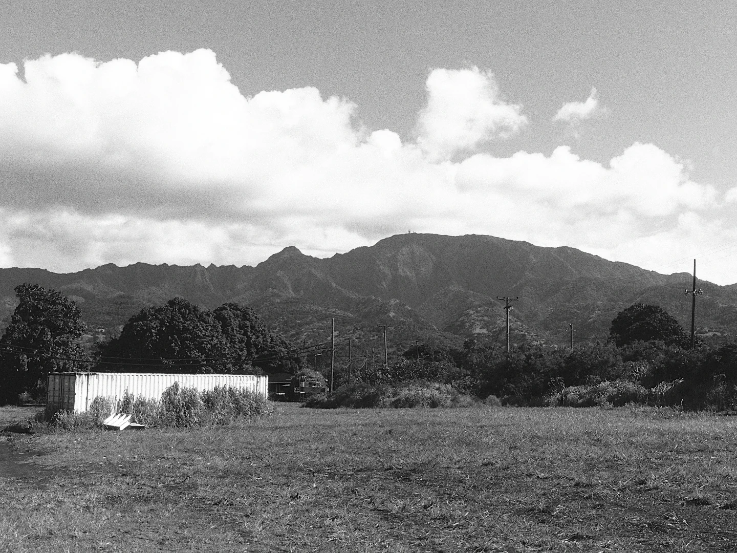 A field below a mountain ridge under white clouds, in black and white with Ilford HP5 Plus 400.
